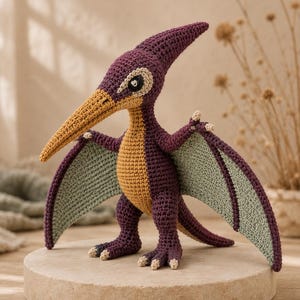 May include: A crocheted pterodactyl toy featuring a deep purple body, a tan underbelly, and a long, pointed mustard yellow beak. The creature has light green wings with purple borders, small claws on its feet and wings, and dark eyes.