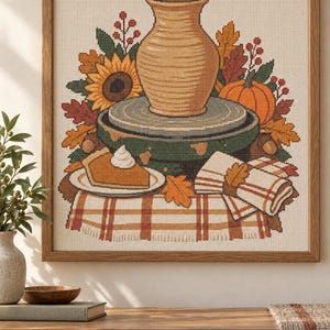 May include: A cross-stitch pattern featuring a tan pottery vase on a green wheel, surrounded by autumn leaves, a sunflower, a pumpkin, and a slice of pumpkin pie with whipped cream on a plaid tablecloth, framed in wood with the text PDF pattern only.