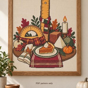 Könnte beinhalten: Ein Kreuzstichmuster in einem Holzrahmen mit einem vertikalen Lineal, einem Winkelmesser, einer brennenden Kerze, einem Stück Kürbiskuchen, einer weißen Pyramide und kleinen Kürbissen. Das Design enthält Herbstblätter, Beeren und karierte Stoffakzente. Text: PDF pattern only.