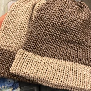 K&ouml;nnte beinhalten: Zwei gestrickte M&uuml;tzen, eine braune und eine beige, mit gerippter Struktur. Die M&uuml;tzen sind zur H&auml;lfte gefaltet und liegen auf einer blau-wei&szlig; gemusterten Oberfl&auml;che.