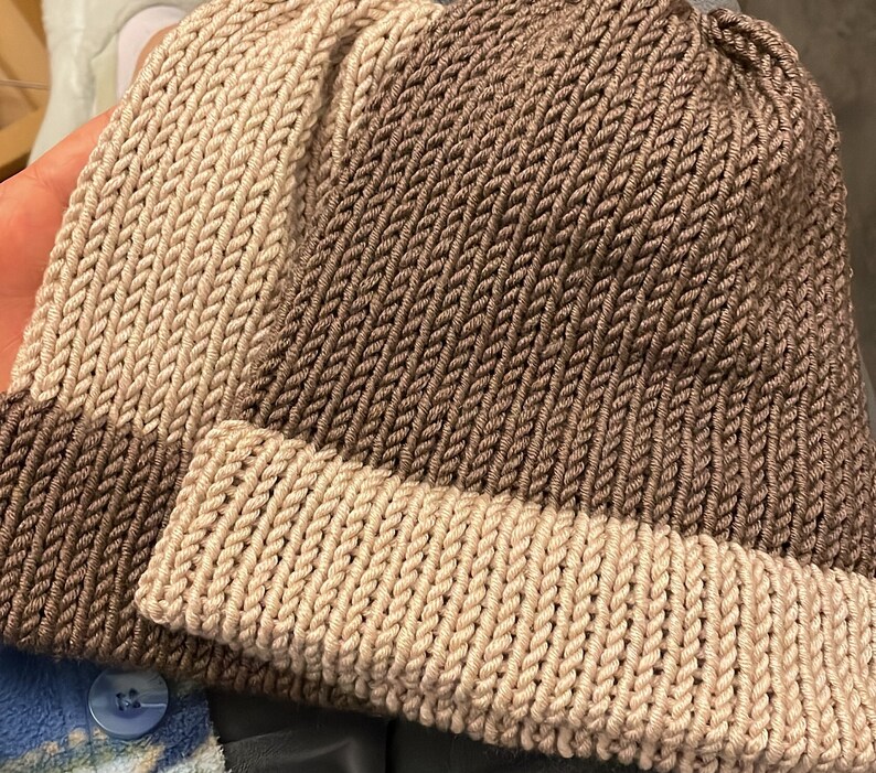 K&ouml;nnte beinhalten: Zwei gestrickte M&uuml;tzen, eine braune und eine beige, mit gerippter Struktur. Die M&uuml;tzen sind zur H&auml;lfte gefaltet und liegen auf einer blau-wei&szlig; gemusterten Oberfl&auml;che.