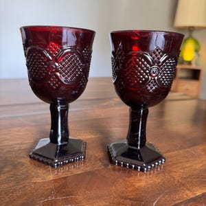 May include: Two ruby red vintage goblets. The glasses have a detailed pattern on the bowl and a hexagonal base. The stem and base are black. The goblets are made of glass and are likely used for serving wine or other beverages.