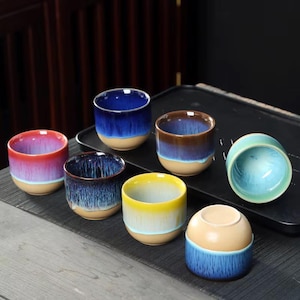 May include: A collection of seven ceramic tea cups featuring a two-tone glaze design. Each cup has a smooth, light beige base and a vibrant, reactive drip-glaze finish in colors including red, deep blue, brown, yellow, and light turquoise.
