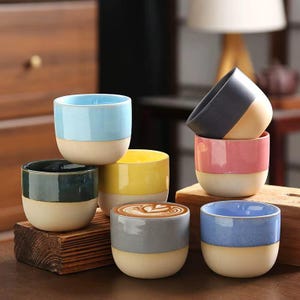 May include: A collection of seven ceramic cups with a two-tone design, featuring a glazed upper section in various colors including light blue, dark green, yellow, gray, pink, and dark blue, with a matte beige base on each cup.