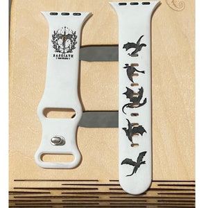 May include: White watch band with black dragon silhouettes and the text "BASGIATH". The band is made of a flexible material and features a silver clasp. The design includes a crest with a sword and wings.