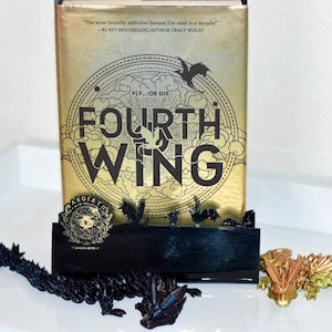 Puede incluir: Un libro dorado con el título "Fourth Wing" y el texto "Fly...or die." El libro está de pie sobre un sujetalibros negro con un diseño de dragón. Hay una figura de dragón negro y una figura de dragón dorada sobre una superficie blanca.