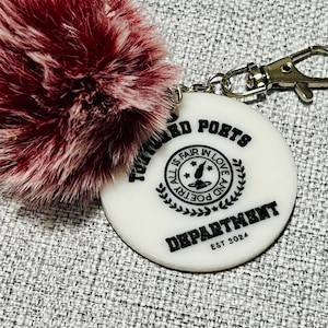 May include: White round keychain with the text "TOWARD POETS DEPARTMENT EST 2024" and a black and white graphic of a quill pen with stars and the text "ALL IS FAIR IN LOVE AND POETRY". The keychain has a fluffy, burgundy pom-pom attached.
