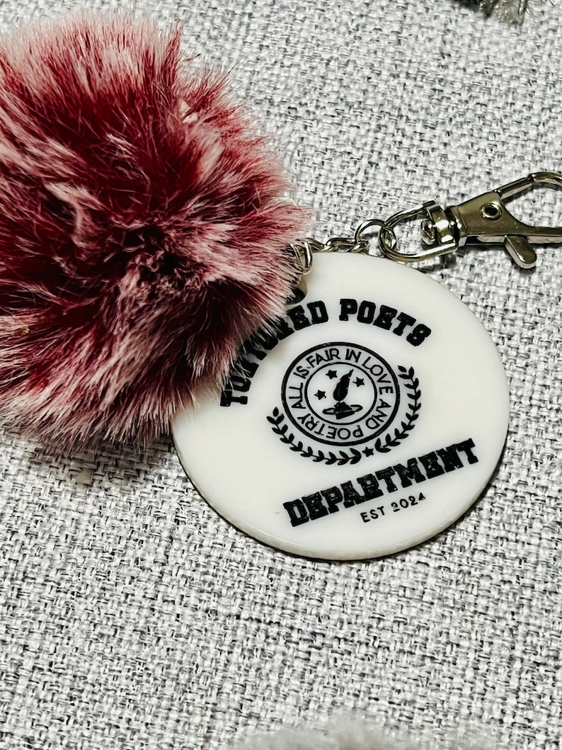 May include: White round keychain with the text "TOWARD POETS DEPARTMENT EST 2024" and a black and white graphic of a quill pen with stars and the text "ALL IS FAIR IN LOVE AND POETRY". The keychain has a fluffy, burgundy pom-pom attached.