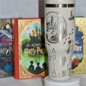 Puede incluir: Un vaso de acero inoxidable blanco con un diseño plateado que presenta el castillo de Hogwarts, el símbolo de las Reliquias de la Muerte y las palabras "Harry Potter". El vaso está sobre un posavasos blanco y tiene una pajita.