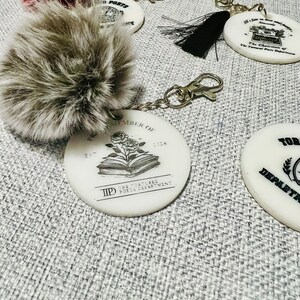 May include: A white ceramic keychain with the text "The Tortured Poets Department" and a book with a rose on top. The keychain is attached to a silver chain and a gray fluffy pom-pom.