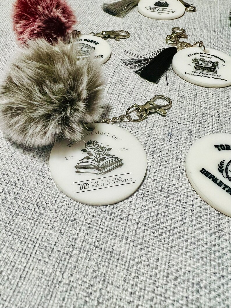 May include: A white ceramic keychain with the text "The Tortured Poets Department" and a book with a rose on top. The keychain is attached to a silver chain and a gray fluffy pom-pom.