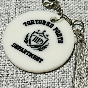 May include: White round keychain with a black and white logo that says "Tortured Poets Department" and a silver tassel.