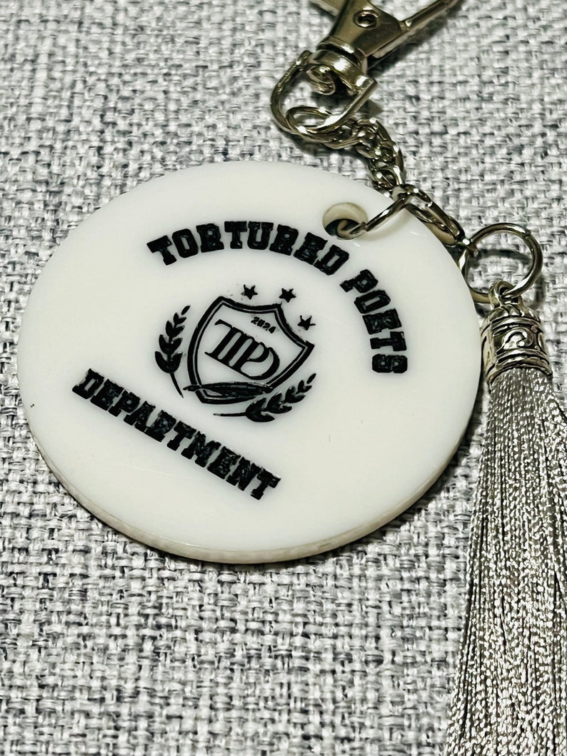 May include: White round keychain with a black and white logo that says "Tortured Poets Department" and a silver tassel.