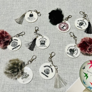 May include: A set of seven white round keychains with black and silver designs. Each keychain features a different design with text that includes "The Tortured Poets Department" and "TIP". The keychains are attached to silver chains and have fluffy pom-poms in various colors, including black, gray, burgundy, and pink.