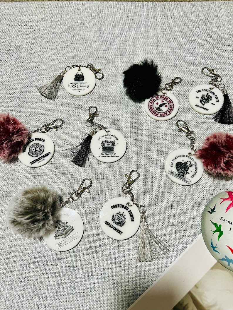May include: A set of seven white round keychains with black and silver designs. Each keychain features a different design with text that includes "The Tortured Poets Department" and "TIP". The keychains are attached to silver chains and have fluffy pom-poms in various colors, including black, gray, burgundy, and pink.