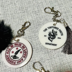 May include: Two white round keychains with black designs and silver key rings. One keychain has a black snake with flowers and the text "The Tortured Poets Est. 2024 Department". The other keychain has a black quill and the text "All is Fair in Love and Poetry".