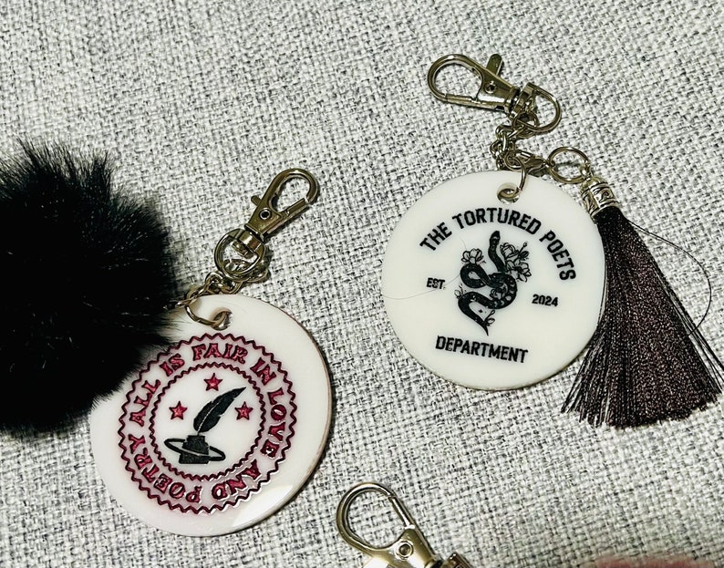 May include: Two white round keychains with black designs and silver key rings. One keychain has a black snake with flowers and the text "The Tortured Poets Est. 2024 Department". The other keychain has a black quill and the text "All is Fair in Love and Poetry".