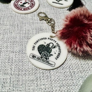 May include: White round keychain with a black and white graphic of a heart with a bomb inside and the text "The Tortured Poets Department" around it. The keychain has a black and white tassel.