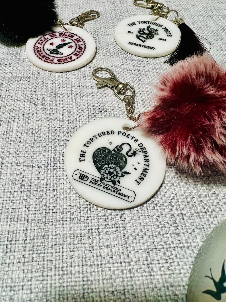 May include: White round keychain with a black and white graphic of a heart with a bomb inside and the text "The Tortured Poets Department" around it. The keychain has a black and white tassel.