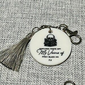 May include: White round keychain with a black and white skull and roses design. The text "Tortured Poets Dept. My Veins of Pitch Black Ink T.S." is printed on the keychain.