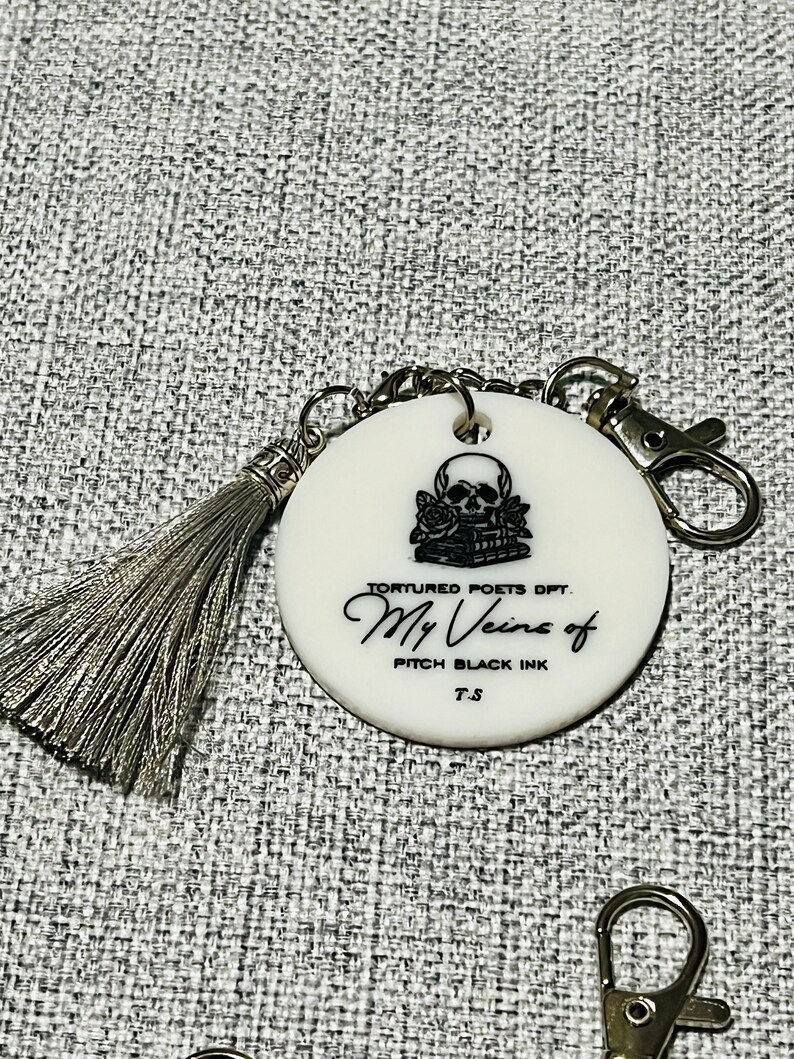 May include: White round keychain with a black and white skull and roses design. The text "Tortured Poets Dept. My Veins of Pitch Black Ink T.S." is printed on the keychain.