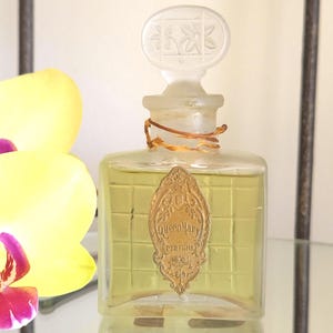 May include: A vintage perfume bottle with a clear glass body and a frosted glass stopper. The bottle is filled with a yellow liquid and features a gold label with the text "Queen Mary Perfume". The stopper is decorated with a floral design.