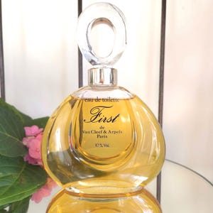 May include: A clear glass perfume bottle with a rounded shape and a clear, oval-shaped stopper. The bottle contains a golden-colored liquid and features the text "First de Van Cleef & Arpels Paris". The bottle is reflected on a mirrored surface.