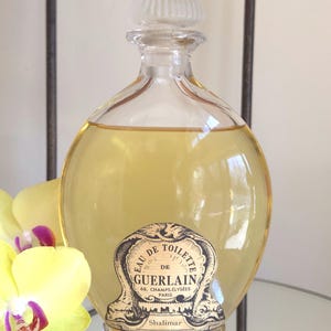 May include: A vintage Guerlain Shalimar Eau de Toilette perfume bottle. The clear glass bottle has a rounded shape and a clear, ridged stopper. The label features the text "Eau de Toilette de Guerlain, 68, Champs-Élysées Paris" and "Shalimar". The perfume inside is a light amber color.