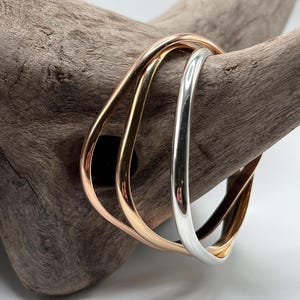 May include: A set of three interlocking bangles featuring a smooth, polished finish. The trio consists of one silver-toned bangle, one yellow gold-toned bangle, and one rose gold-toned bangle, each with a rounded, slightly irregular, wavy shape.