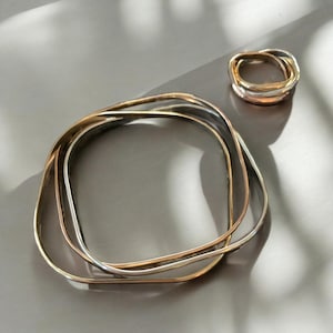 May include: Three silver bangles stacked on top of each other, forming a square shape. The bangles are made of different widths and have a polished finish.