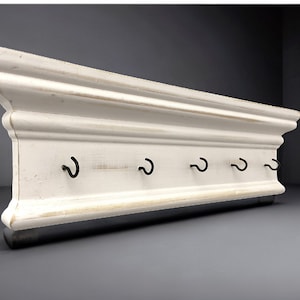 May include: White wooden wall mounted key rack with five black metal hooks. The rack has a decorative, ornate design.