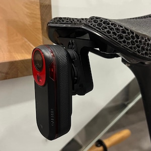 Puede incluir: Una cámara de ciclismo Garmin negra con una luz roja y una lente unida a un poste de asiento de bicicleta negro.