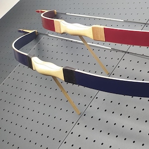May include: Two recurve bows, one red and one blue, are displayed against a gray pegboard. The bows have wooden handles and limbs, with the red bow above the blue bow. The bowstrings are silver.