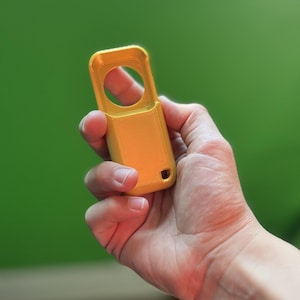 May include: A bright yellow, rectangular object with a circular opening and a small hole at the bottom. The object is held in a hand against a green background. The object appears to be 3D printed.