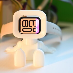 May include: White stand with a white Apple Watch with a pink and black watch face. The watch is in a white stand that looks like a robot.