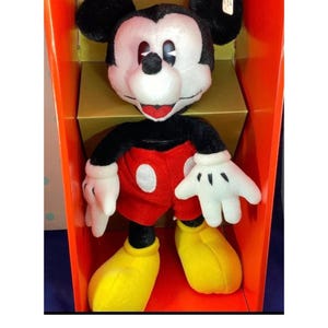May include: A plush Mickey Mouse doll, featuring a black head with large ears, a white face, and a red mouth. The doll wears a black top, red shorts, white gloves, and yellow shoes. It is displayed inside an orange box.
