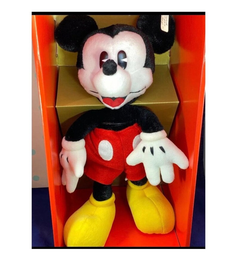 May include: A plush Mickey Mouse doll, featuring a black head with large ears, a white face, and a red mouth. The doll wears a black top, red shorts, white gloves, and yellow shoes. It is displayed inside an orange box.