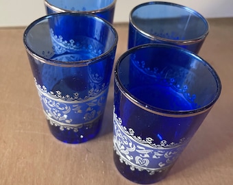 Può includere: Quattro bicchieri blu cobalto con bordo argentato e motivo decorativo bianco e argento. I bicchieri sono disposti su una superficie marrone chiaro.
