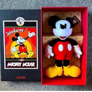 May include: A Mickey Mouse plush toy in a red box. The plush has a black head and ears, white face and hands, a red body with two white buttons, and yellow shoes. The box has a black lid with a Mickey Mouse graphic and the text "Introducing Mickey Mouse".