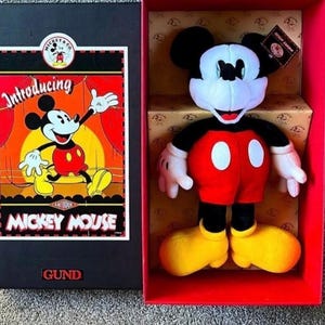 May include: A Mickey Mouse plush toy in a red box. The plush has a black head and ears, white face and hands, red shorts with white buttons, and yellow shoes. The box features a vintage Mickey Mouse graphic and the text "Introducing Mickey Mouse".