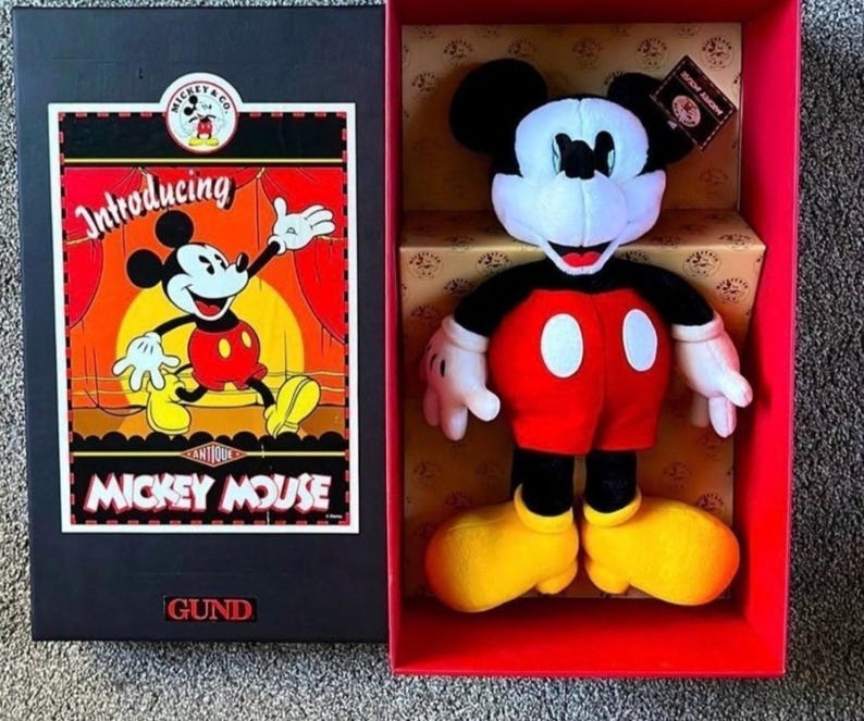 May include: A Mickey Mouse plush toy in a red box. The plush has a black head and ears, white face and hands, red shorts with white buttons, and yellow shoes. The box features a vintage Mickey Mouse graphic and the text "Introducing Mickey Mouse".