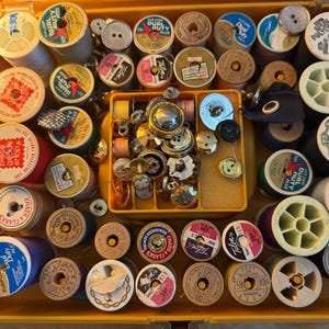 Puede incluir: Una caja de costura amarilla llena de carretes de hilo de varios colores y tamaños, junto con una variedad de botones. Los carretes tienen etiquetas con textos como "Dual Duty" y "Talon 100% Polyester".