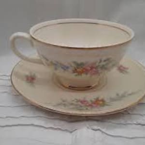 May include: Cream-colored teacup and saucer set with floral designs in pink, blue, and yellow. The cup has a curved handle and sits on a matching saucer, both with gold trim. The set is likely made of porcelain.