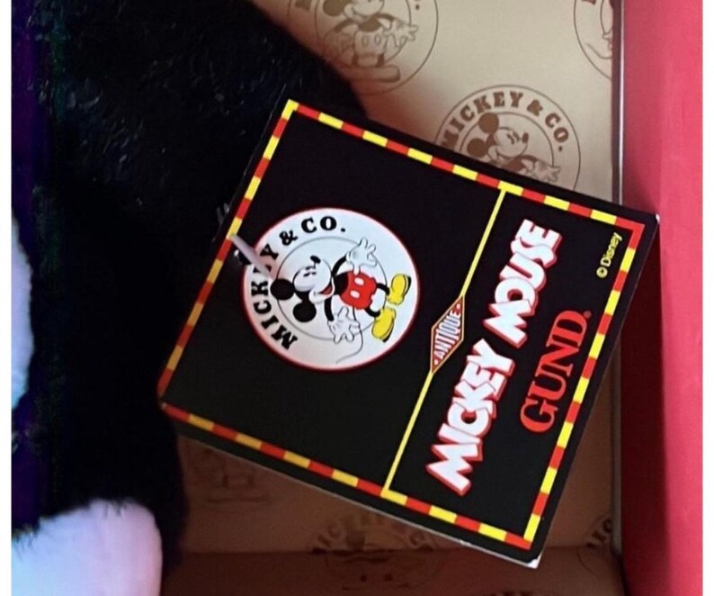 May include: A black tag with red and yellow trim features the text "Mickey Mouse Gund Antique" and a Mickey Mouse graphic. The tag is attached to a black and white plush toy.