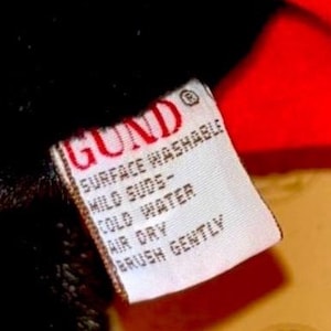 May include: Close-up of a white tag with red text and a red border. The tag reads "GUND" at the top, followed by care instructions: "SURFACE WASHABLE, MILD SUDS, COLD WATER, AIR DRY, BRUSH GENTLY". The tag is attached to a black plush material.