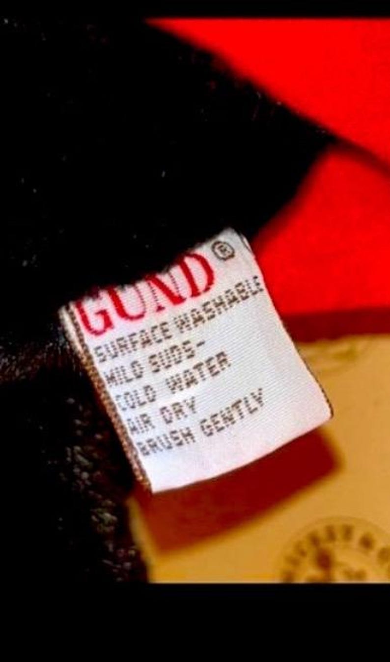May include: Close-up of a white tag with red text and a red border. The tag reads "GUND" at the top, followed by care instructions: "SURFACE WASHABLE, MILD SUDS, COLD WATER, AIR DRY, BRUSH GENTLY". The tag is attached to a black plush material.
