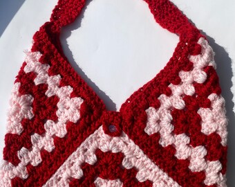 Puede incluir: Una bolsa de mano de crochet roja y rosa con un cierre de botón único. La bolsa tiene una base ancha y plana y una correa larga.
