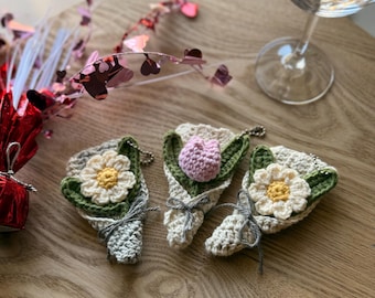 May include: Three crocheted flower bouquets with a green stem and leaves. One bouquet features a pink crocheted tulip, while the other two feature white crocheted daisies with yellow centers. The bouquets are tied with twine and have a small metal ring attached.