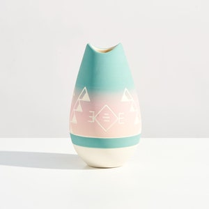 Può includere: Un vaso in ceramica blu chiaro e rosa con un motivo geometrico. Il vaso ha la forma di una lacrima e una base bianca.