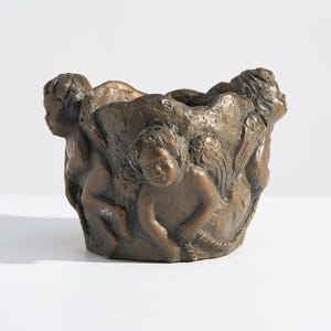 Könnte beinhalten: Bronze-farbener Pflanztopf mit einem skulpturalen Design von cherubischen Figuren. Das Gefäß hat eine abgerundete Form, wobei die Figuren anscheinend den Rand stützen. Die Cherubim sind in verschiedenen Posen, was dem Stück Dynamik verleiht. Geeignet für kleine Pflanzen.